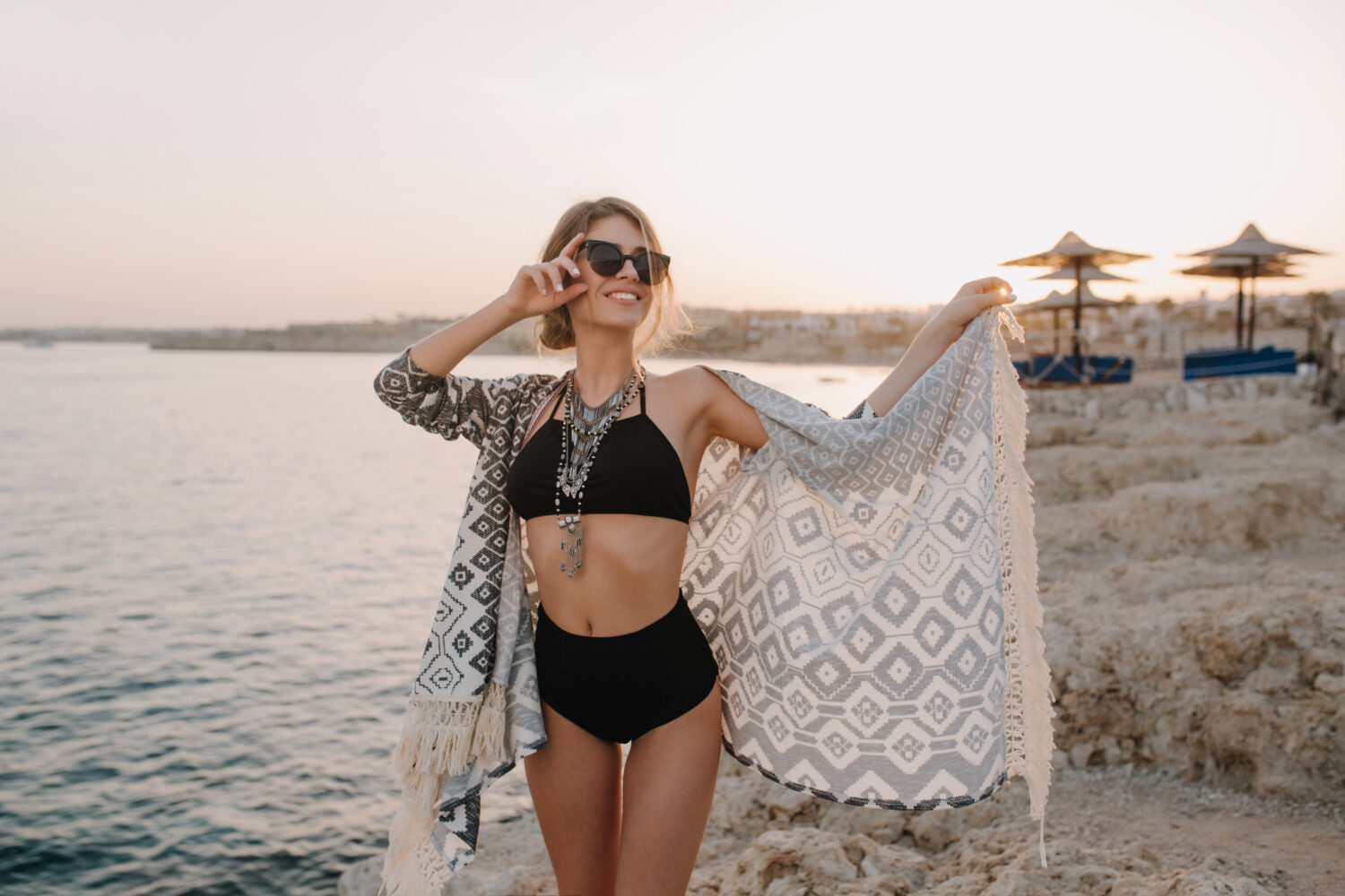 Fasionable beautiful girl posing on beach, on sunset. Sexy slim woman wearing black bikini, swimsuit with hight waist, cardigan, cape with ornaments, beautiful sea, rock on background..